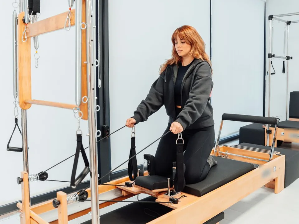 パーカーと黒いレギンスの女性が、タワー付きのピラティスのマシンの上でひざをつき、両手でベルトを引いている