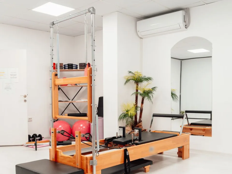白い部屋に、タワー付きのピラティスのマシンとピンクのボール、観葉植物が置かれたスタジオ